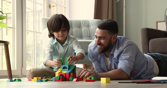 Inspired junior school age boy play on warm wooden floor by picture window together with caring father. Young dad little son spend free time for hobby activity build castle from bright colored details