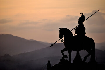 Silhueta de cavaleiro com armadura, chapéu com pena e uma lança - topo de um monte com montanhas...