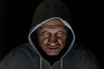 A man in a hood, a lighted face on a dark background. Evil grimace, low key.