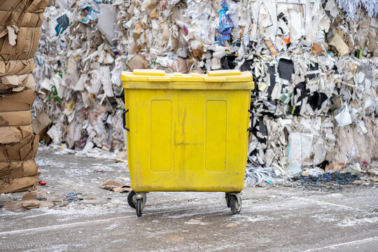 Photo of a yellow dumpster with bales of waste paper and rubbish in the background. Pile of pressed waste paper bales in the yard.