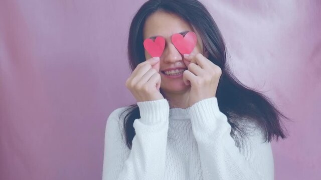 Smiling funny young brunette in a white sweater smiles, blinks and waving red hearts on a pink background. Valentine's Day