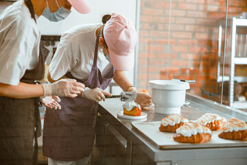 Confectioner trainee spreads cream onto sponge cake while colleague teaches her in bakery