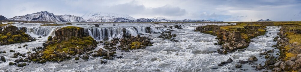 Season changing in southern Highlands of Iceland. Picturesque waterfal Tungnaarfellsfoss panoramic autumn view.  Landmannalaugar mountains under snow cover in far.