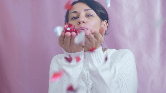 Smiling funny young brunette in a white sweater sends air kisses blowing on red hearts on a pink background. Valentine's Day