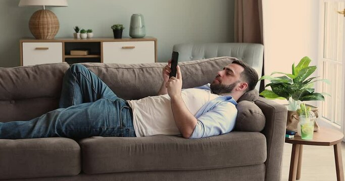 Relaxed millennial guy lie on back on soft comfortable couch at living room spend lazy weekend watching video on mobile phone screen. Calm young man rest use cellphone to check email read news online