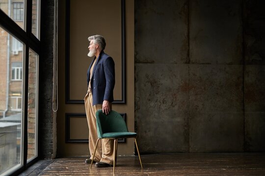 Full length shot of pensive grey haired middle aged man in casual wear looking out a large window while standing in modern loft interior
