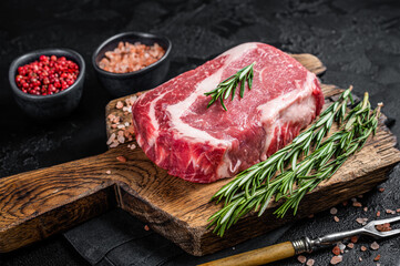 Raw Rib-eye Steak, beef marbled meat on wooden board with rosemary. Black background. Top view