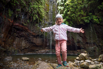 Traveler girl wearing sport jacket and raising hands at waterfall. Travel lifestyle. View from back.
