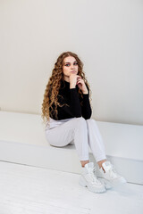 A beautiful woman with long curly hair in a black sweater, white pants, white leather sneakers. New collection of women's casual shoes