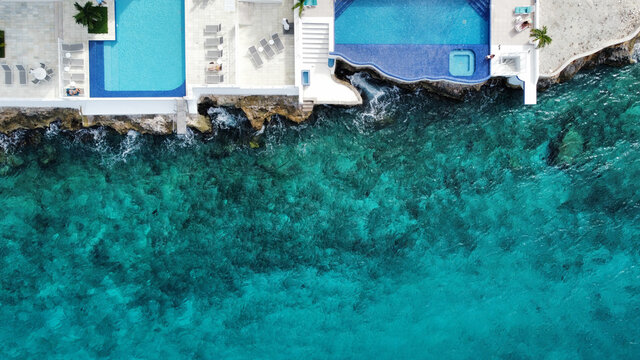 Clear, turquoise ocean water from the Caribbean sea meets the rocky shore of Cozumel, Mexico with vibrant blue swimming pools. Aerial view captured by drone.