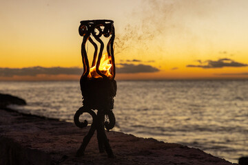 Fire in a Tiki torch burns on a tropical island beach next to the Caribbean sea during sunset. 