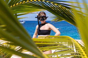 Young woman wearing snorkel and mask on a tropical island beach framed by palm tree fronds. 