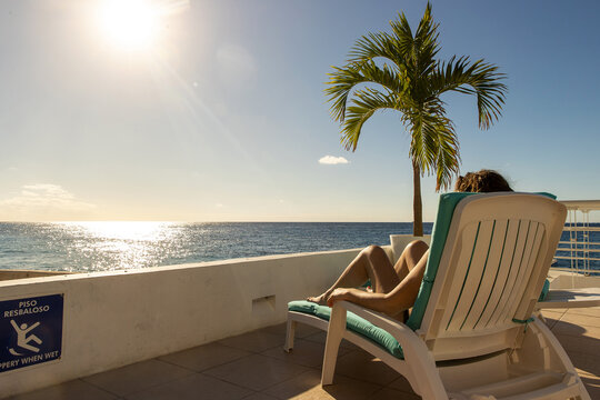 Woman relaxing poolside on vacation getting a tan under the Caribbean sun with a dramatic view of the ocean from her chair on the tropical island of Cozumel in Mexico. 