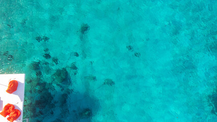 A white dock with vibrant red umbrella and furniture is surrounded by turquoise blue Caribbean sea water. The ocean is clear and coral reefs can be seen below. Aerial vew by drone. 