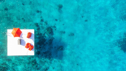 A white dock with vibrant red umbrella and furniture is surrounded by turquoise blue Caribbean sea water. The ocean is clear and coral reefs can be seen below. Aerial vew by drone. 