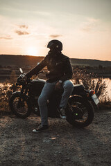 Portrait of confident motorcyclist man in motorcycle helmet. Young driver biker looking away outdoors alone near river. Ready for trip. Cafe racers, motorbike aesthetics and vintage design concept