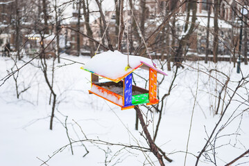 A bright multicolored bird feeder hanging outdoors in winter against the background of a gray forest, covered with snow.