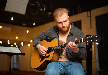 A young guy with a beard plays an acoustic guitar in a room with warm lighting