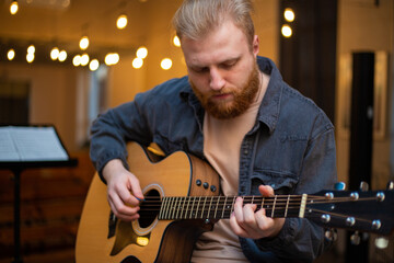 Obraz premium A young guy with a beard plays an acoustic guitar in a room with warm lighting