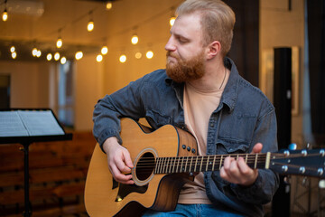 Obraz premium A young guy with a beard plays an acoustic guitar in a room with warm lighting