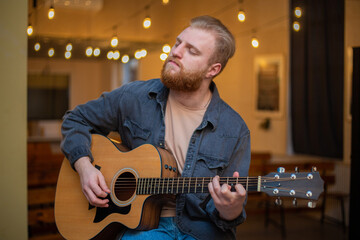 Obraz premium A young guy with a beard plays an acoustic guitar in a room with warm lighting