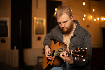 A young guy with a beard plays an acoustic guitar in a room with warm lighting