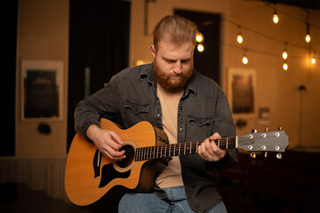 Obraz premium A young guy with a beard plays an acoustic guitar in a room with warm lighting