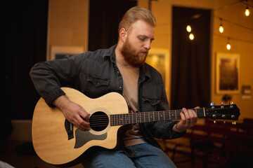 A young guy with a beard plays an acoustic guitar in a room with warm lighting