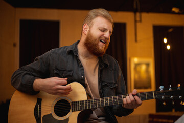 Obraz premium A young guy with a beard plays an acoustic guitar in a room with warm lighting