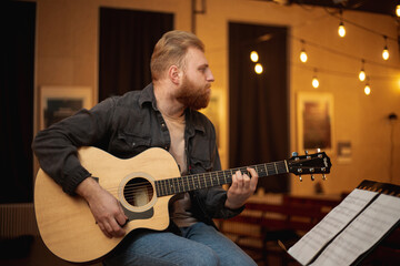 Obraz premium A young guy with a beard plays an acoustic guitar in a room with warm lighting