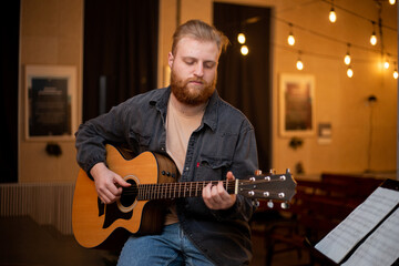 Obraz premium A young guy with a beard plays an acoustic guitar in a room with warm lighting