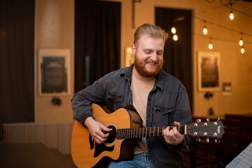 Obraz premium A young guy with a beard plays an acoustic guitar in a room with warm lighting