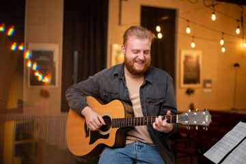 A young guy with a beard plays an acoustic guitar in a room with warm lighting