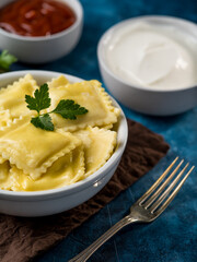 Close-up. Ravioli in a white earthenware bowl with parsley leaf and butter on a brown napkin, fork. In the background are sauces. Blue background. Restaurant, hotel, cafe, home cooking.