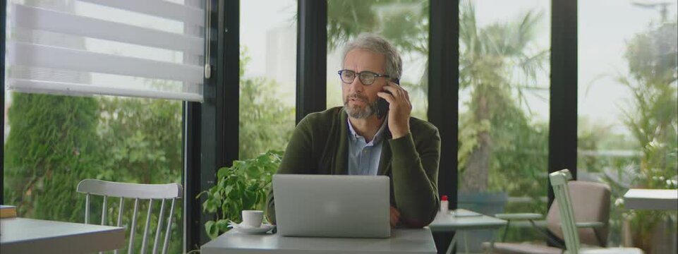 Mature man with serious face expression calling business partner. Sitting at glass wall in cafe. Entrepreneur working outside office.