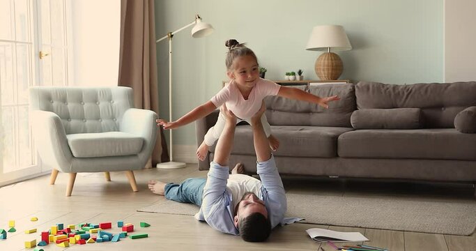 Cute little girl spread arms like wings enjoy freedom in imaginary flight balance in air on strong dad hands. Loving daddy lie on floor with underheating help small daughter imitate plane raise kid up