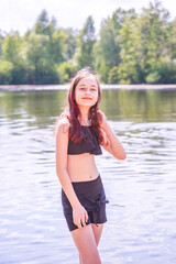 A teenage girl by the river in a black swimsuit and black shorts in summer. Children and rest.