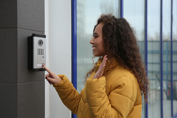 Fototapeta na wymiar Young African-American woman ringing intercom while waving to camera near building entrance
