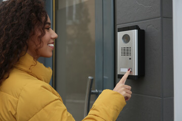 Young African-American woman ringing intercom with camera near building entrance - obrazy, fototapety, plakaty