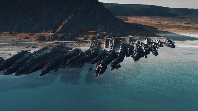 Aerial view of the long jagged reefs on the Varanger shores. Waves slowly roll over the dark rocks. narrow road following the shoreline. Slow-motion, pan right