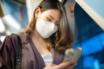 Asian businesswoman with protective face mask using smart phone and standing at window while commuting by train. Lifestyle portrait of a people holding with smart phone at the modern train