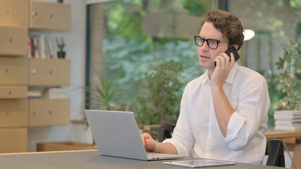 Young Man Talking on Smartphone while Using Laptop in Modern Office