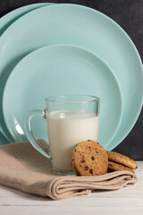 A glass mug with milk and oatmeal cookies on a white wooden table against a background of plates leaning against the wall.
