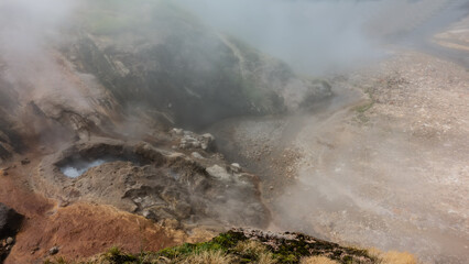Fototapeta na wymiar Geyser cauldron on the hillside. Boiling water foams. Hot steam hides the surroundings. A river flows through a rocky bed from thermal springs. Kamchatka. Valley of Geysers