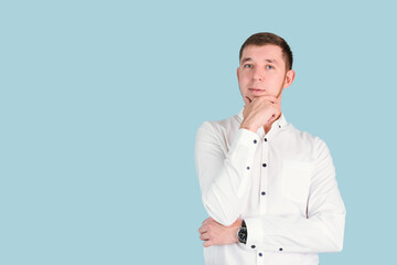 A thoughtful bearded man in a white shirt holds his hand on his chin while standing on a blue background, copyspace