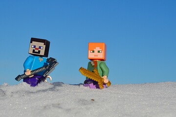 Obraz premium LEGO Minecraft figures of Steve with iron pickaxe and Alex with golden sword standing knee deep in real snow sunny winter day, blue skies in background. 