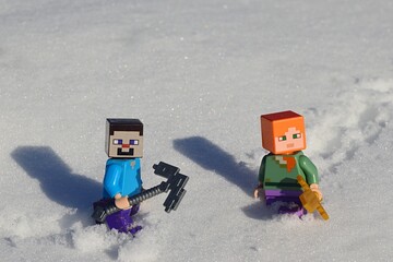 Obraz premium LEGO Minecraft figures of Steve with iron pickaxe and Alex with golden sword walking through deep real snow, sunny winter day,.