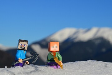 Obraz premium LEGO Minecraft figure of Steve with iron pickaxe and Alex with golden sword, standing knee deep in real snow, snowy mountain and blue skies in background. 