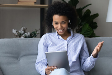 Smiling pretty african american woman in earphones looking at digital tablet screen, holding distant web camera video call conversation, talking speaking communicating distantly, virtual event concept