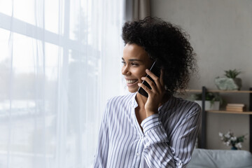 Dreamy happy young african american biracial woman looking out of window, involved in pleasant cellphone call conversation, talking chatting with friends, speaking communicating remotely with family.